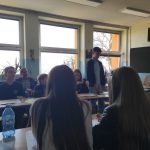 Grupa uczniów siedzi przy ławkach w klasie, niektórzy zwróceni twarzą do przodu, a inni zwróceni w stronę ucznia, który stoi i mówi. Dorośli siedzą przy oknach w tle. Sala jest jasno oświetlona światłem dziennym.