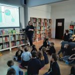 Kobieta wygłasza prezentację dla grupy dzieci siedzących na podłodze w bibliotece. Ekran projekcyjny za nią pokazuje rysunki postaci z kreskówek i wirusa. Półki z książkami ustawione są wzdłuż ścian.