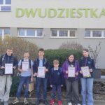 Sześciu uczniów stoi przed budynkiem z napisem DWUDZIESTKĄ, trzymają certyfikaty i się uśmiechają. Za nimi widać krzewy i okna, a w prawym dolnym rogu znajduje się małe logo szkoły.