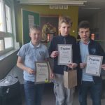 Trzech chłopców stoi w pomieszczeniu, trzymając certyfikaty. Są zwróceni twarzą do kamery i się uśmiechają. W tle widać okno, zielone ściany i obraz. Chłopcy noszą jasnoniebieskie koszule z kołnierzykami i ciemnym emblematem.