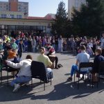 Grupa osób siedzących i grających na instrumentach muzycznych na zewnątrz. Otacza ich tłum stojących gapiów. Sceneria wydaje się być szkołą lub ośrodkiem społecznościowym z drzewami i budynkiem w tle.