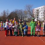 Grupa dzieci i kilku dorosłych ubranych w kolorowe, różnorodne kostiumy, w tym zwierzęta, klauni i postacie, stoi na czerwonej bieżni sportowej. W tle widać apartamenty i nagie drzewa pod czystym, błękitnym niebem.