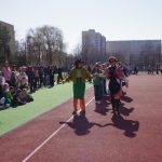 Grupa ludzi ustawia się na boisku sportowym pod czystym niebem. Dwie osoby w kolorowych kostiumach stoją na początku kolejki. Dzieci i dorośli obserwują z boku, z drzewami i budynkami w tle.