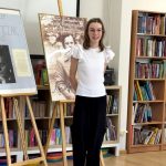 Osoba w białym topie i czarnych spodniach stoi uśmiechnięta przed regałami z książkami i tablicami wystawowymi ze zdjęciami i tekstem. Miejsce wydaje się być biblioteką lub księgarnią.