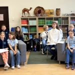 Grupa dzieci i nastolatków ze szkoły, niektórzy siedzą na sofach, a inni na krzesłach, pozują w bibliotece lub klasie. Półki wypełnione książkami i przedmiotami dekoracyjnymi ustawione są wzdłuż tylnej ściany.