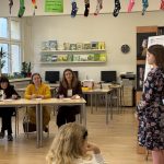 Młoda dziewczyna w kwiecistej sukience stoi przed klasą i prezentuje. Trzy kobiety siedzą przy stole i ją obserwują. W tle widać regały z książkami, komputery i kolorowe skarpetki zwisające z sufitu.