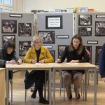 Trzy kobiety siedzą przy stole i piszą, a za nimi na tablicy wiszą czarno-białe zdjęcia. Chłopiec w szkolnym mundurku przechodzi obok po prawej stronie. Sceneria wydaje się być klasą lub biblioteką z ekspozycjami edukacyjnymi.