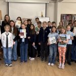 Grupa dzieci i kilku dorosłych stoi razem w bibliotece, trzymając książki i certyfikaty, uśmiechając się do kamery. Półki na książki ustawione są wzdłuż ścian, a w tle widać ekran projektora.