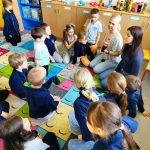 Grupa małych dzieci i dwóch dorosłych siedzi w kręgu na kolorowym dywanie z uśmiechniętą buźką w klasie, angażując się w dyskusję lub aktywność. W tle znajdują się półki z książkami i butelkami.