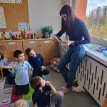 Kobieta stoi przy oknie w klasie, pokazując coś grupie małych dzieci siedzących i klęczących na kolorowym dywanie, wszyscy uważnie ją obserwujący. W tle widoczne są plakaty edukacyjne i rośliny.