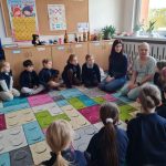 Grupa małych dzieci i dwóch dorosłych siedzi w kręgu na kolorowym dywanie w klasie, zaangażowani w dyskusję. W tle widoczne są dekoracje klasy i plakaty edukacyjne.