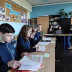 Czterech uczniów siedzi przy biurkach w klasie, czytając i zaznaczając prace. Dwóch dorosłych i inny uczeń pracują na laptopach w tle. W pokoju są niebieskie ściany i tablica ogłoszeń ze zdjęciami i KLASA EUROPEJSKA.