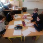 Czwórka dzieci siedzi przy stole w klasie, pisząc na papierach. W pomieszczeniu znajdują się materiały edukacyjne, tablica i laptop w pobliżu. Uczniowie intensywnie skupiają się na swojej pracy, a na stole leżą różne artykuły piśmiennicze.