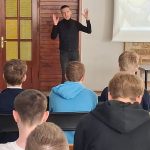 Osoba w czarnym stroju wygłasza prezentację przed publicznością w klasie. Gestykuluje rękami, podczas gdy projekcja wyświetla tekst na ścianie. Publiczność siedzi na krzesłach, zwrócona twarzą do mówcy.