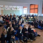 Grupa dzieci i kilku dorosłych siedzi w klasie, oglądając prezentację. Niektóre dzieci siedzą na matach, a inne na krzesłach. Ściany zdobią półki i prace uczniów, a okna mają pomarańczowe żaluzje.