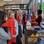 Wolontariusze w pomarańczowych kamizelkach zebrali się przy stole w sklepie, rozmawiając z ludźmi i udzielając informacji. Stół jest zaopatrzony w różne artykuły spożywcze, a w tle widoczne są znaki sklepu.