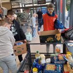 Trzech młodych ludzi pakuje produkty spożywcze, takie jak makaron, olej i słoiki, do pudełek przy kasie w sklepie spożywczym. Osoba w tle korzysta z wózka sklepowego. W tle widoczne są półki i szyldy sklepowe.