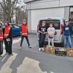 Grupa dzieci stoi na chodniku obok zaparkowanego vana. Niektóre z nich noszą czerwone kamizelki. Na ziemi leżą dwa pudełka z jedzeniem. W tle widać drzewa i zaparkowane samochody.