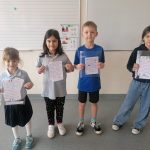 Czwórka dzieci stoi w klasie trzymając kolorowe, ręcznie pisane papiery. Są ubrane swobodnie i stoją przed tablicą z nutami. Na podłodze znajduje się emblemat szkoły. Wyglądają na dumnych ze swojej pracy.