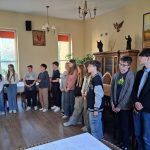 Grupa młodych ludzi stoi w rzędzie w pokoju z drewnianymi meblami. Ściany zdobią oprawione obrazy i krucyfiks. Niektórzy trzymają papier, a wszyscy wydają się być uważni.