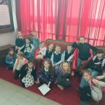 Grupa małych dzieci siedzi i klęczy na czerwonym dywanie przed czerwonymi zasłonami, pozując do grupowego zdjęcia. Większość się uśmiecha, niektórzy robią znaki pokoju. Noszą swobodne i półformalne ubrania z plecakami.