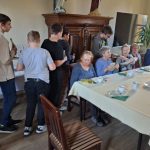 Grupa ludzi zebrała się wokół stołu, niektórzy siedzą, a niektórzy stoją. Siedzące osoby, głównie starsi dorośli, trzymają książki i szklanki. Stół jest nakryty do posiłku kubkami i talerzami. W tle widać regał z książkami i wysoką roślinę.