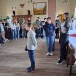 Grupa dzieci i nastolatków stoi w pokoju z drewnianymi podłogami i dużymi oknami. Niektórzy z nich zebrali się w kącie, podczas gdy dwie dziewczyny stoją w środku. Wzdłuż jednej ściany znajdują się stoły, a przestrzeń zdobią obrazy i rośliny.