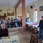 Grupa ludzi, w tym studenci i starszy mężczyzna, zebrali się w pokoju. Starszy mężczyzna siedzi, a młoda osoba stoi i mówi. Stoły są nakryte do posiłku z filiżankami do herbaty i talerzami. Ściany zdobią obrazy i dekoracje. Naturalne światło wypełnia pokój.