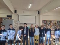 Grupa małych dzieci stoi w klasie, uśmiechając się do kamery. Są ubrani w pasujące do siebie mundurki szkolne. W tle w pomieszczeniu znajdują się regały wypełnione książkami, a na ścianie ekran projekcyjny.