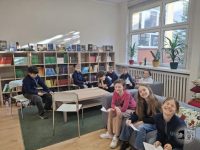 Grupa dzieci siedzi w bibliotece, niektóre na krzesłach, inne na podłodze, w tle regały z książkami i okna. Uśmiechają się i trzymają papiery, a w pobliżu okien stoją doniczkowe rośliny.