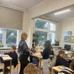 Nauczyciel stoi w klasie, wchodząc w interakcję z uczniami siedzącymi przy ławkach. Są oni zaangażowani w czytanie i dyskusję. Książki są wystawione na półkach, a na biurku stoi komputer. Duże okna wpuszczają naturalne światło z zewnątrz.