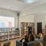 Osoba stoi w klasie i wygłasza prezentację. Za nią ekran wyświetla tekst i obrazy. Kilku uczniów siedzi i uważnie obserwuje. Ściany są ustawione na półkach wypełnionych książkami.