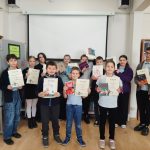 Grupa dzieci stoi w bibliotece, trzymając dyplomy i książki, uśmiechając się do kamery. Są ubrani swobodnie i ustawieni przed białym ekranem, z półkami na książki po obu stronach.