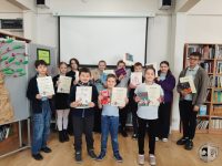 Grupa dzieci stoi w bibliotece, trzymając dyplomy i książki, uśmiechając się do kamery. Są ubrani swobodnie i ustawieni przed białym ekranem, z półkami na książki po obu stronach.