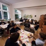 Dzieci siedzą przy stołach w klasie i pracują nad projektami artystycznymi, w tle widać regały z książkami.