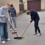 Trzech młodych ludzi zamiata brukowaną powierzchnię miotłami na zewnątrz. Dwóch z nich ma na sobie bluzy z kapturem i dżinsy, a trzeci kurtkę puchową i spodnie dresowe. W tle widać drzwi garażowe i odpływ.