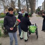 Dwóch młodych ludzi pomaga starszej kobiecie poruszającej się na wózku inwalidzkim na chodniku. Jeden trzyma bukiet róż. Inna osoba, częściowo widoczna, robi zdjęcie telefonem. W tle widać drzewa i domy.