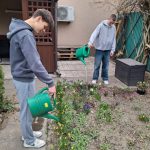 Dwie osoby podlewają ogród zielonymi konewkami. Stoją na wybrukowanym terenie obok klombu z różnymi kwitnącymi roślinami. W tle widać budynek i drewnianą konstrukcję.