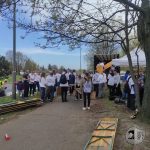 Grupa ludzi zbiera się na zewnątrz, niektórzy trzymają drewniane ramy. Inni stoją w pobliżu białego namiotu i zaparkowanej ciężarówki. Jest pogodny dzień, a wzdłuż wybrukowanego chodnika rosną drzewa. Kilka osób nosi pasujące do siebie koszulki, co sugeruje skoordynowane wydarzenie lub aktywność.