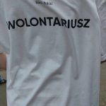 Na zdjęciu widać osobę ubraną w biały t-shirt. Na plecach widnieje napis WOLONTARIUSZ w kolorze czarnym, a także logo i nazwa banku, Bank Polski.