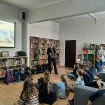 Osoba wygłasza prezentację przed studentami siedzącymi w bibliotece z regałami na książki i ekranem.