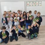 Grupa uśmiechniętych dzieci w klasie trzyma doniczkowe rośliny, pozując razem przed tablicą. Niektóre dzieci klęczą z przodu, podczas gdy inne stoją za nimi, dumnie prezentując swoje rośliny.