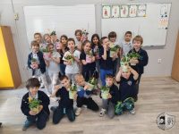 Grupa uśmiechniętych dzieci w klasie trzyma doniczkowe rośliny, pozując razem przed tablicą. Niektóre dzieci klęczą z przodu, podczas gdy inne stoją za nimi, dumnie prezentując swoje rośliny.