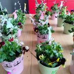 Małe pastelowe doniczki wypełnione zielonymi roślinami, bratkami i gałązkami wierzby stoją na drewnianym stole. Niektóre doniczki mają wycięte papierowe dekoracje, takie jak motyle i króliki. Czerwone szafki są w tle.