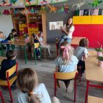 Kobieta czyta książkę z obrazkami grupie małych dzieci w kolorowej klasie udekorowanej rękodziełem i roślinami. Dzieci siedzą przy stołach, słuchają i obserwują czytelnika.