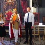 Trzy osoby stoją w środku; centralna postać nosi koronę i pelerynę, trzyma berło, podczas gdy inni stoją po obu stronach. W tle widać duże królewskie tło i plakaty.