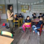 Nauczyciel stoi przed młodymi uczniami siedzącymi w ławkach w klasie, niektórzy z nich mają podniesione ręce.