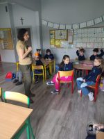 Nauczyciel stoi przed młodymi uczniami siedzącymi w ławkach w klasie, niektórzy z nich mają podniesione ręce.