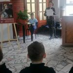 Kobieta czyta na mównicy, podczas gdy uczeń słucha obok; pozostali siedzą naprzeciwko nich w jasnym pomieszczeniu.