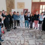 Grupa dzieci stoi w rzędzie w pomieszczeniu zamkniętym, trzymając ręce przy ustach, a dorosły je obserwuje.